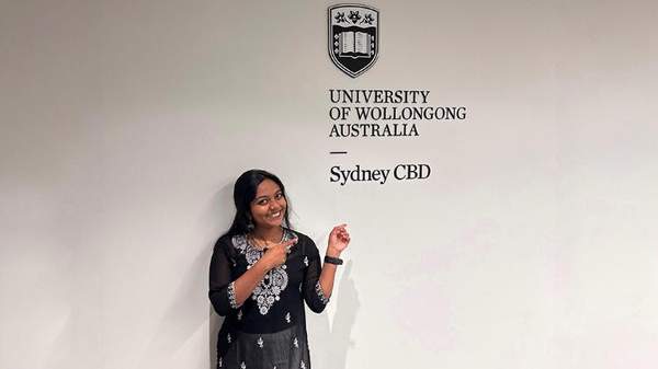 Sydney Business School, UOW student, Gopika Radha smiles as she points to the Sydney Business School, UOW logo on a white wall behind her.
