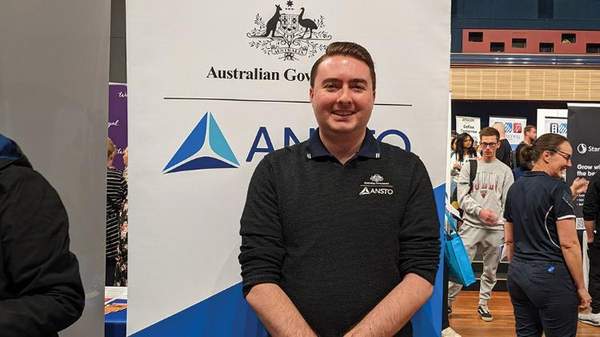 Patrick WIlson stands infront of a floorbanner. His company, ANSTO is printed on the banner. He stands with his hands together infront of him