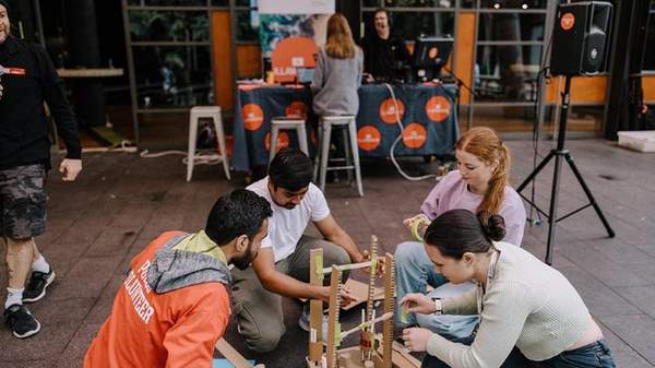 Four people sitting outside on a paved area building a structure with various items. One people is holding a wheel of tape.