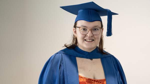 ýapp graduate Catie Brown wears a blue graduation cap and stands against a white wall. She is smiling at the camera. Photo: Paul Jones