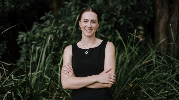 Denelle Cosier stands with her arms crossed, in a black dress, in front of a garden background. Photo: Michael Gray