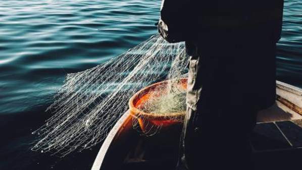 Fishing nets are being pulled into a boat from the ocean. The silhouette of a person is visible.
