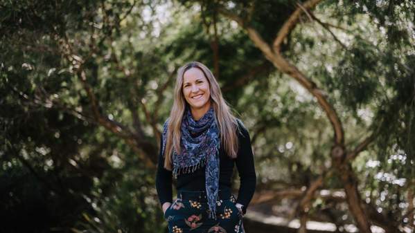 Dr Haidee Cadd stands with her hands in her pockets in front of a background of trees. She is smiling and wears a black top and black shirt with a wattle pattern. Photo: Michael Gray