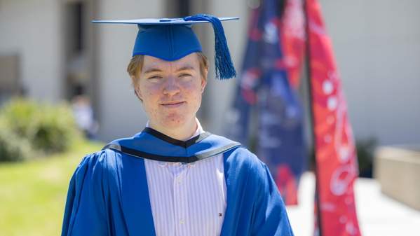 Graduating student Jackson Gunn stands in front of the Sports Hub, wearing a blue cap and gown. Photo: Mark Newsham