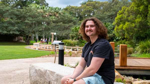 UOW student Luke Bradbery, wearing a black shirt and blue shorts, sits with the ý campus in the background. Photo: Adam Mansfield