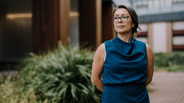 A woman stands outside a building, which looms over her in the background. She wears glasses and a dark blue dress. Photo: Michael Gray