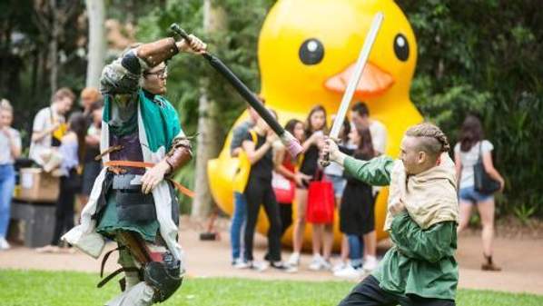 Two students take part in a pretend sword fight while a group of students watch on. The big UOW duck is in the background. Photo: Paul Jones