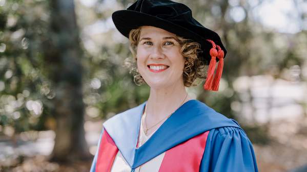 Dr Natalie Day wears a graduation cap and gown and stands in front of green trees on campus. Photo: Michael Gray
