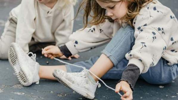 A young child sits on the ground while tying up shoelaces