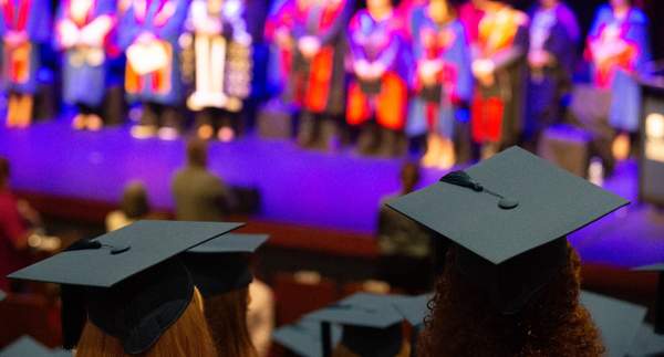 A shot of students' graduation caps with people standing on stage in the background. Photo: Paul Jones