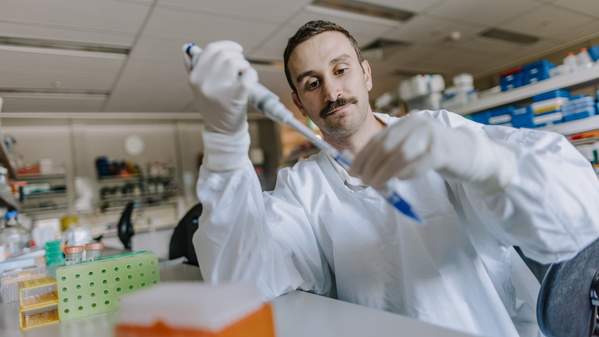 A man wearing a white lab coat holds a syringe as he sits at a science lab bench. Photo: Michael Gray