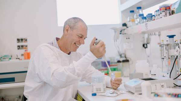 A man sits at a laboratory desk, wearing a white coat and looking at a microscope. Photo: Michael Gray