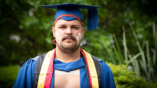 UOW graduate Zachary Stewart, wearing graduation gowns with the Indigenous colours and his traditional dress, stands against a green bush background. Photo: Paul Jones