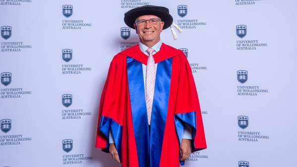 Professor Joe Chicharo stands against a UOW backdrop, wearing a blue and red graduation gown and cap. Photo: Andy Zakeli