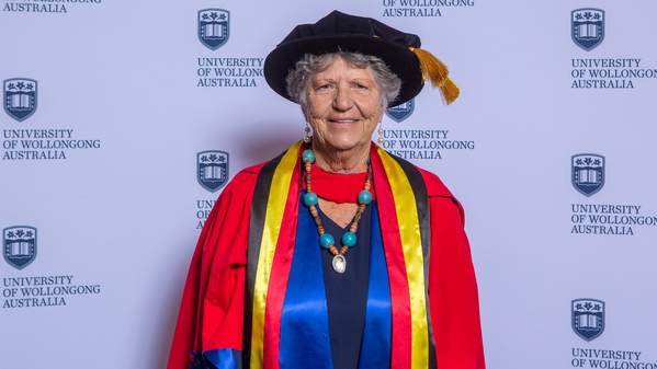 Aunty Joyce Donovan is smiling and standing in front of a UOW media wall wearing a graduation gown and cap. Photo: Andy Zakeli