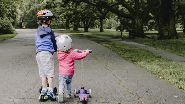 Two young children ride their scooters on a leafy path. Photo: Unsplash