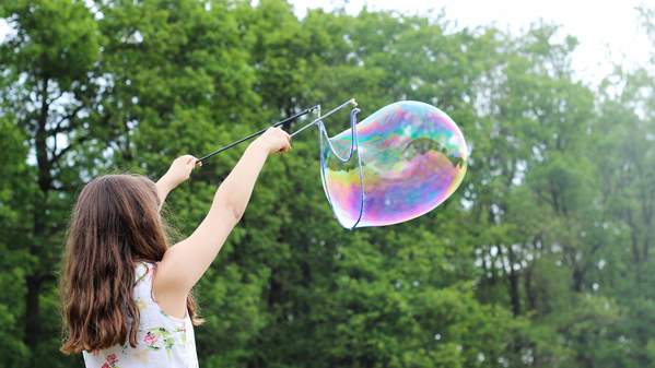 A girl with brown hair and her back to the camera creates a large rainbow bubble. She is in the outdoors. Photo: Unsplash