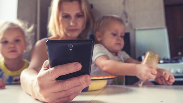 A woman looks at her phone while holding on to the two small children. Photo: Vitola Klein/Unsplash
