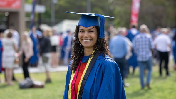 Janaya Pender, wearing a red dress, blue and red graduation gown, and blue cap, smiles at the camera. There are people gathered in the background. Photo: Mark Newsham