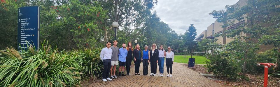 Group of people standing on a paved path in a semi circle with a green leafy trees behind them