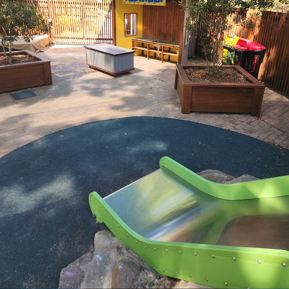 froma childs point of view down, looking down the green slied, a mud kitchen is seen toward the boundary fence