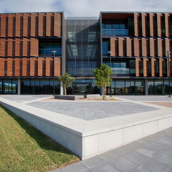 Modern building with a glass facade and wooden slats, featuring a courtyard with two small trees and paved walkways, under a partly cloudy sky.