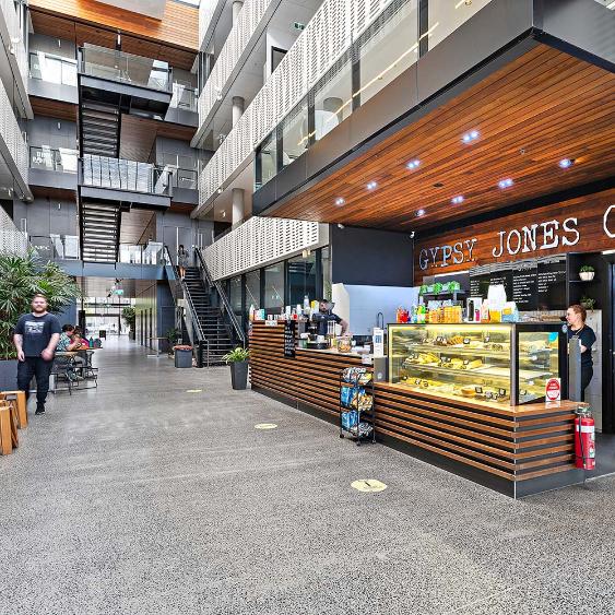A spacious modern café area featuring a wooden counter, glass display case, and staircases in a light-filled atrium setting.