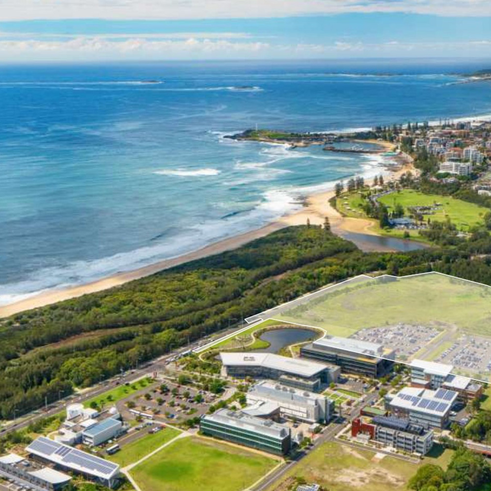 Aerial view of a coastal city with a beach, green spaces, and several modern buildings with parking lots near the shoreline.