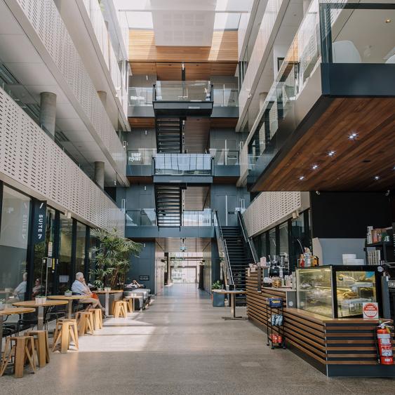 Modern atrium with glass railings, central staircase, seating area with people, and a café counter on the right, under bright skylight.
