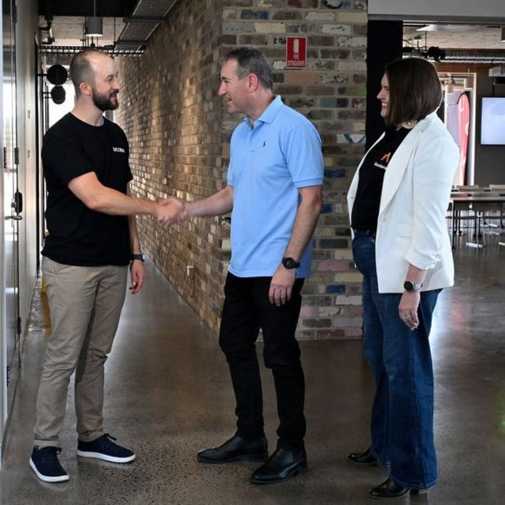 Two men shake hands in a modern office space, while a woman observes, dressed in casual business attire. Brick walls and tables in background.