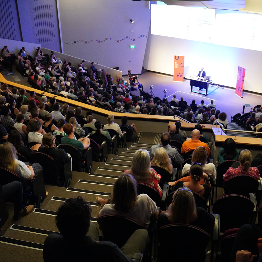 looking toward the stage from the rear of the theatre. You can see a presenter on stage and the room filled with guests