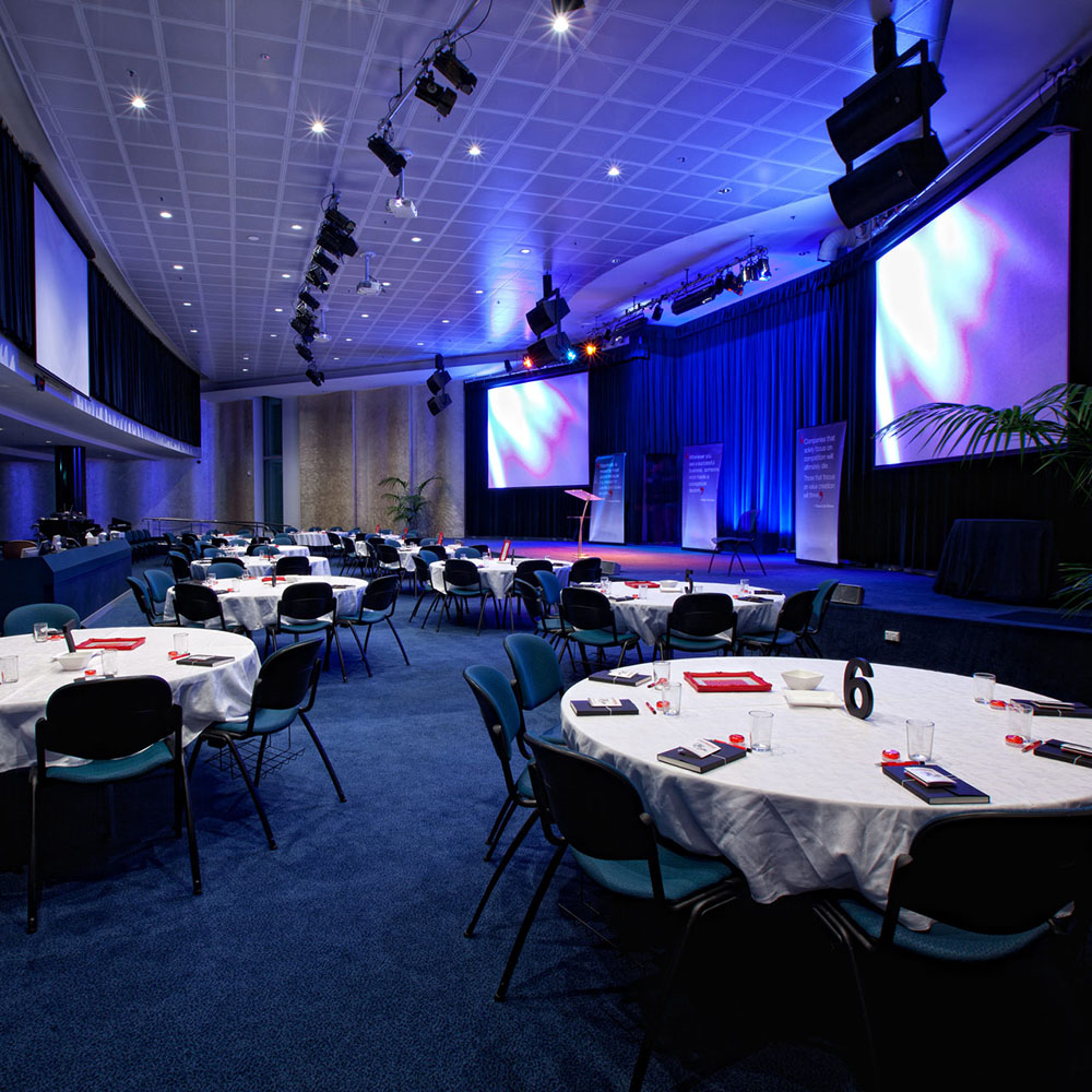 Conference room with round tables and chairs on a blue carpet. Two large screens and a podium are on a stage with blue lighting, creating a formal atmosphere.
