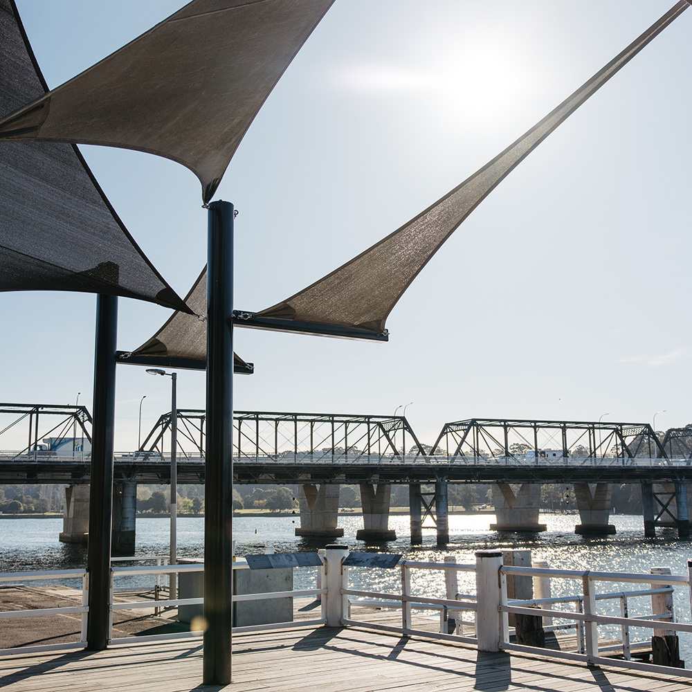 Sunlight shines over a waterfront scene featuring modern shade sails and a bridge reflected in the water below.