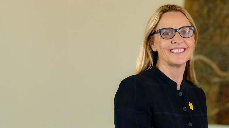 Tanya-Buchanan-smiles-at-the-camera.-She-wers-a-black-button-up-blazer-and-is-wearing-a-daffodil-day-pin