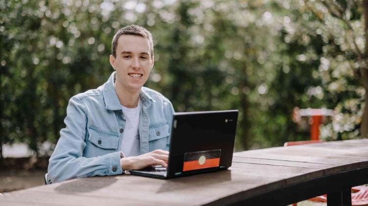 A male presenting student sitting at a bench outside, looking at the camera happily while a laptop sits in front of them