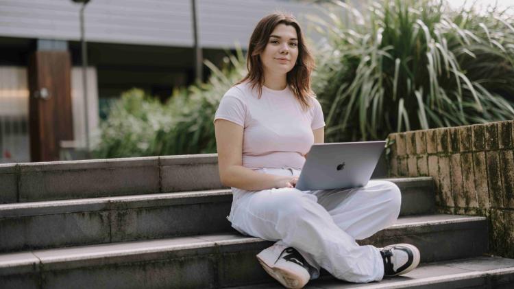 A female presenting student sitting on outside stairs with a laptop in their lap smiling at the camera