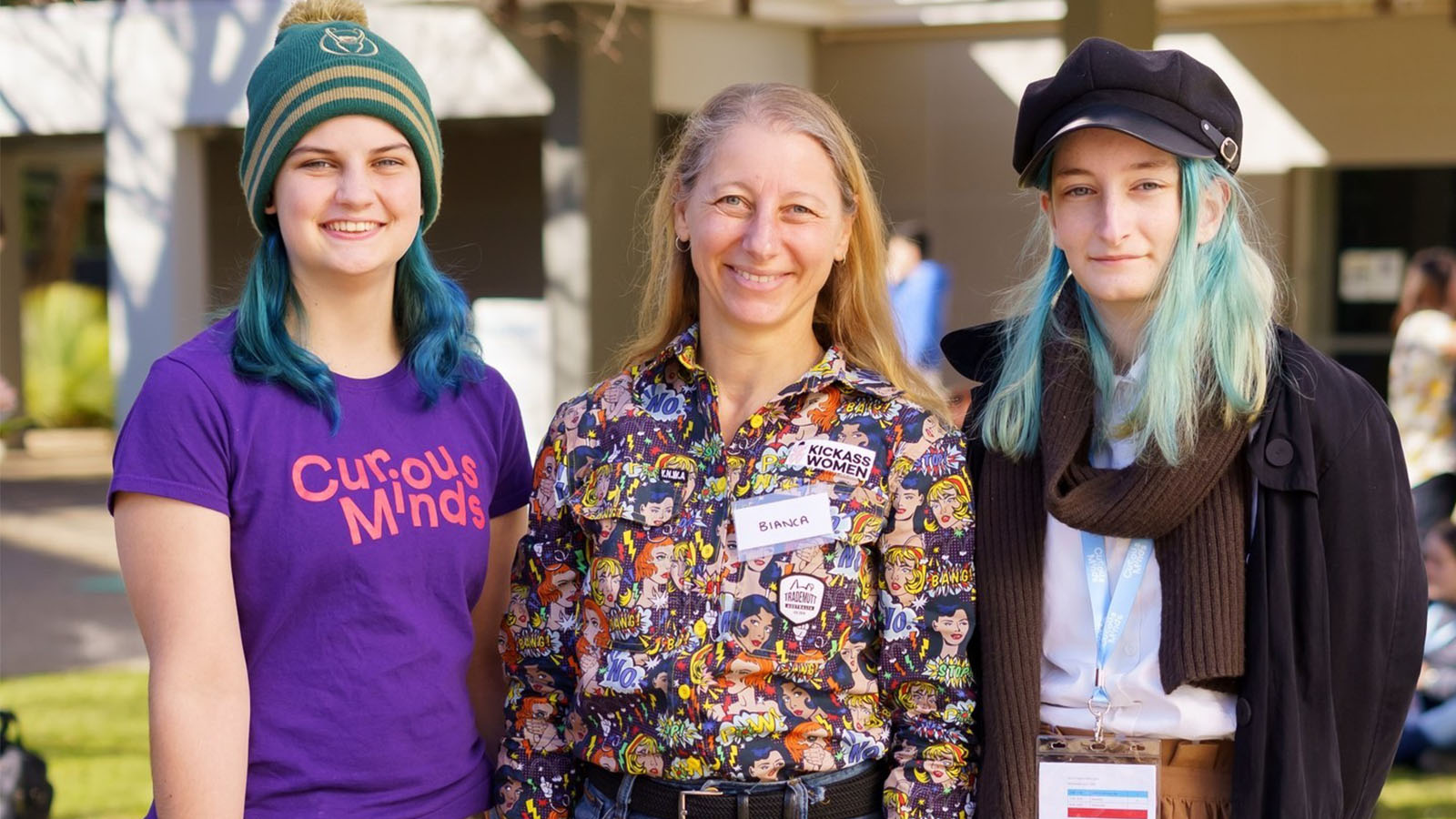 Bianca Shepherd, wearing a colourful comic-print shirt with a 'Kickass Women' badge, stands smiling between two young women with blue hair at an outdoor event. One wears a 'Curious Minds' t-shirt, and the other wears a black coat and hat.
