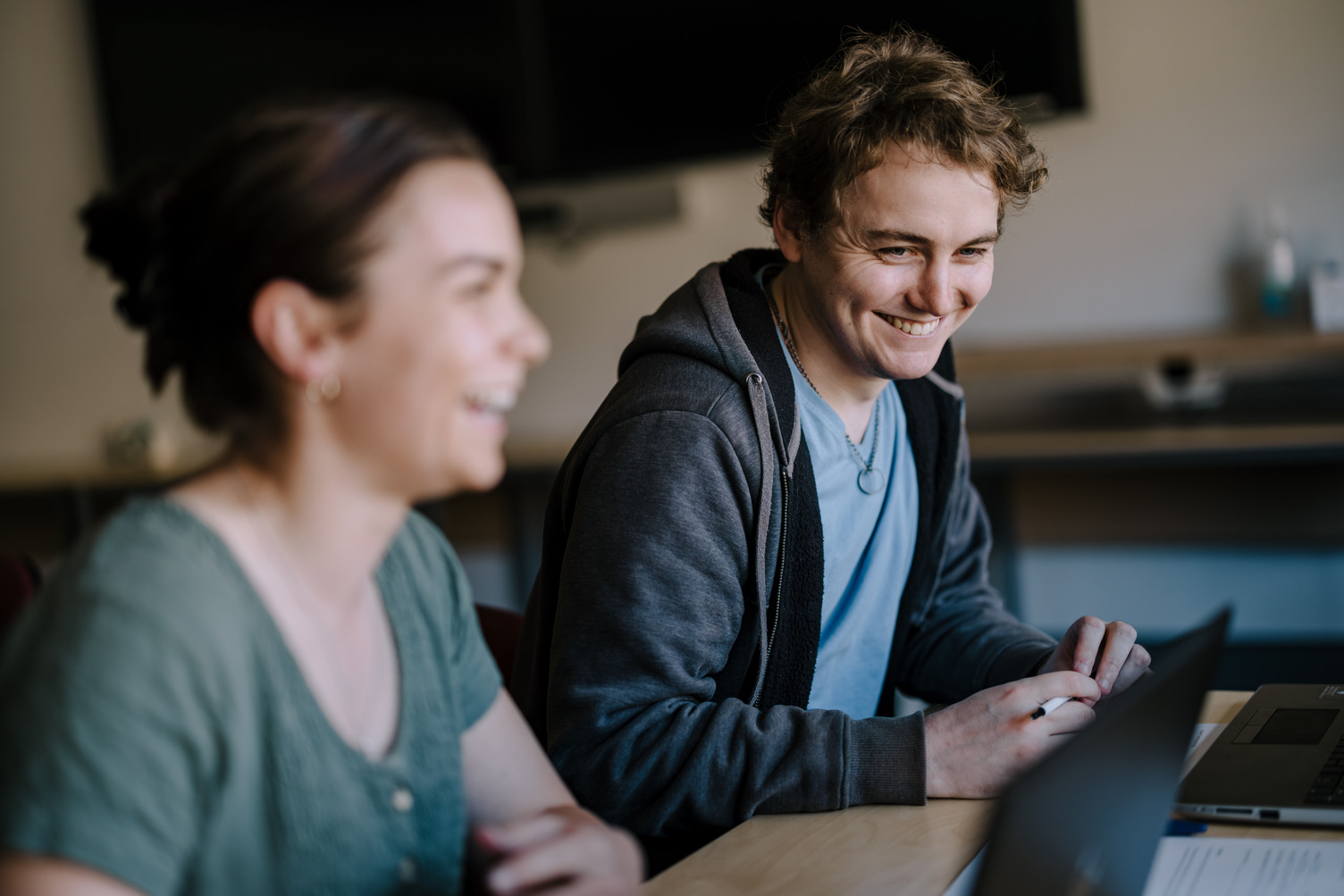 A male student in focus and a female student out of focus sitting at a desk, both laughing.