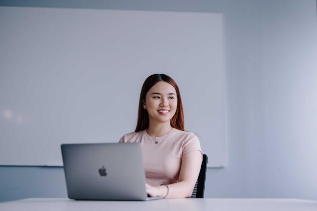 A female presenting student sitting at a white desk with a Mac laptop, smiling and looking off to the side