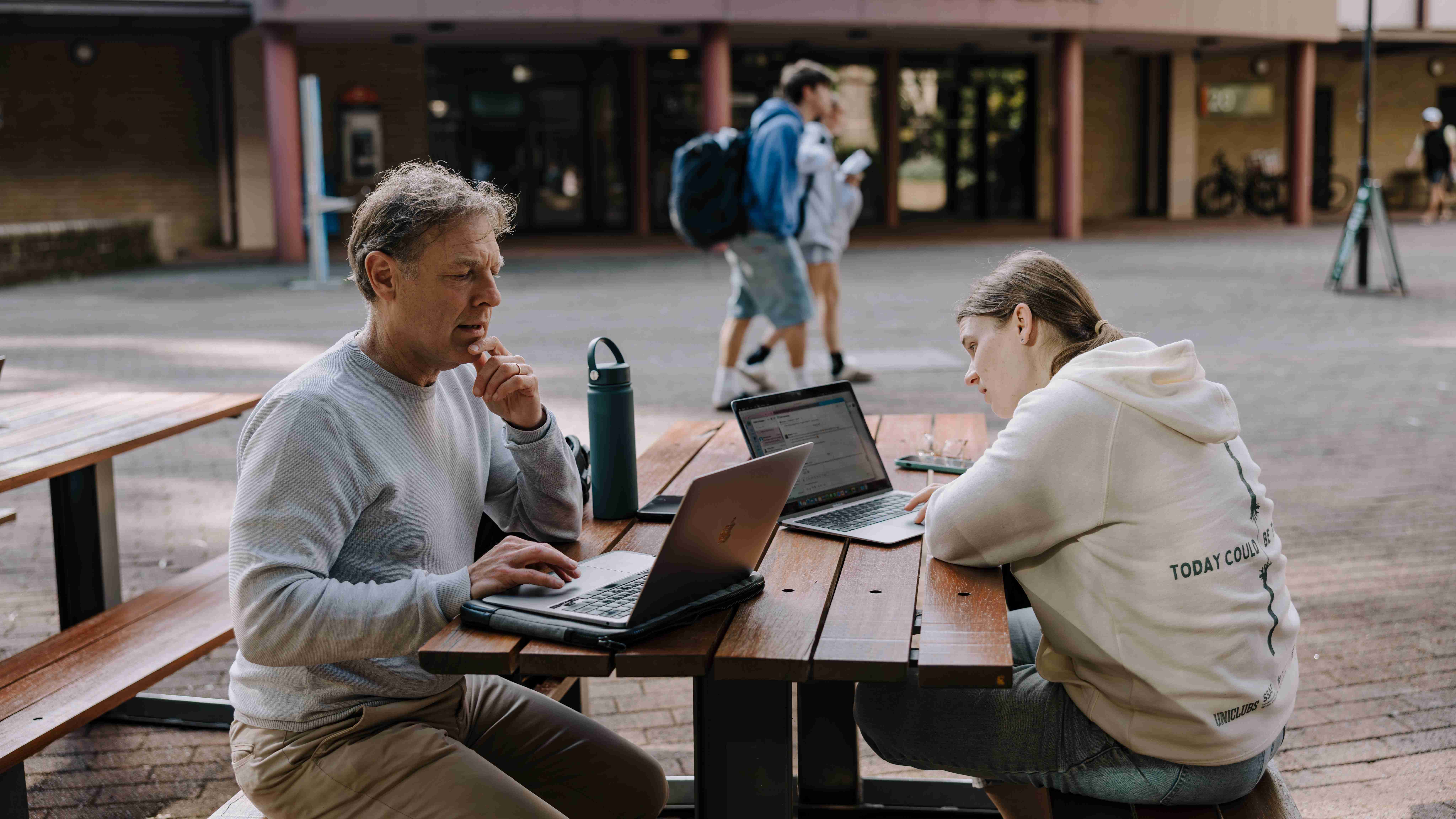 Two people working on laptops at an outdoor wooden table, with a green water bottle between them.