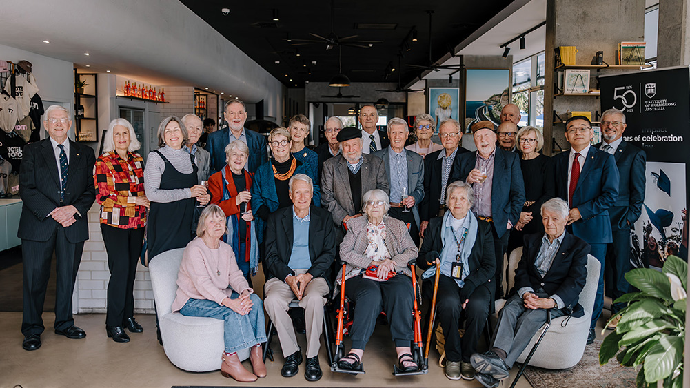 A diverse group of elderly people, dressed in formal attire, gather in a modern, well-lit room. They pose together, smiling warmly, conveying a sense of camaraderie and celebration.