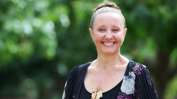 Faye McMillan is standing against a wood panel wall, smiling. She wears a white blazer and frameless glasses