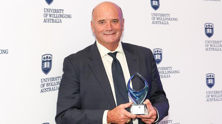 Paul Smith is wearing a black business shirt and is in front of a black wall. The image is in black and white