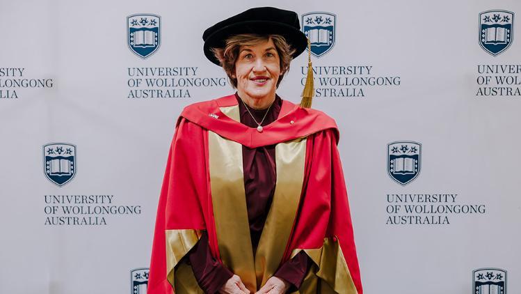 Christine McLoughlin AM in red Honorary Gown in front of a white wall with repeated UOW logo