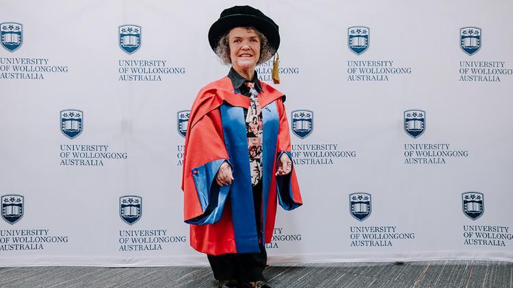 Dr Debra Keenahan in red Honorary Gown in front of a white wall with repeated UOW logo