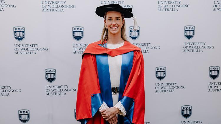 Emma McKeon AM  in red Honorary Gown in front of a white wall with repeated UOW logo