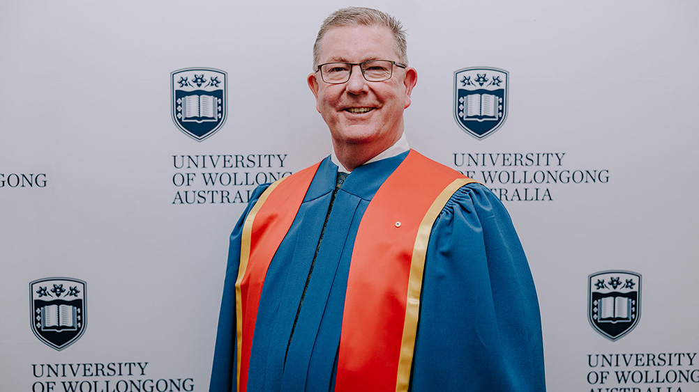 Graham Lancaster AM in blue Honorary Gown in front of a white wall with repeated UOW logo