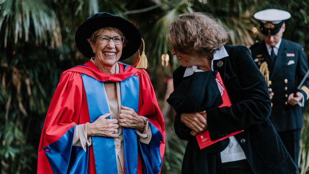 Candid outdoor photo of Her Excellency the Honourable Margaret Beazley AC KC (left) laughing while wearing red and blue academic regalia and a black mortarboard with a gold tassel. She stands next to a woman in a black velvet blazer holding a red folder, with a naval officer visible in the blurred background.