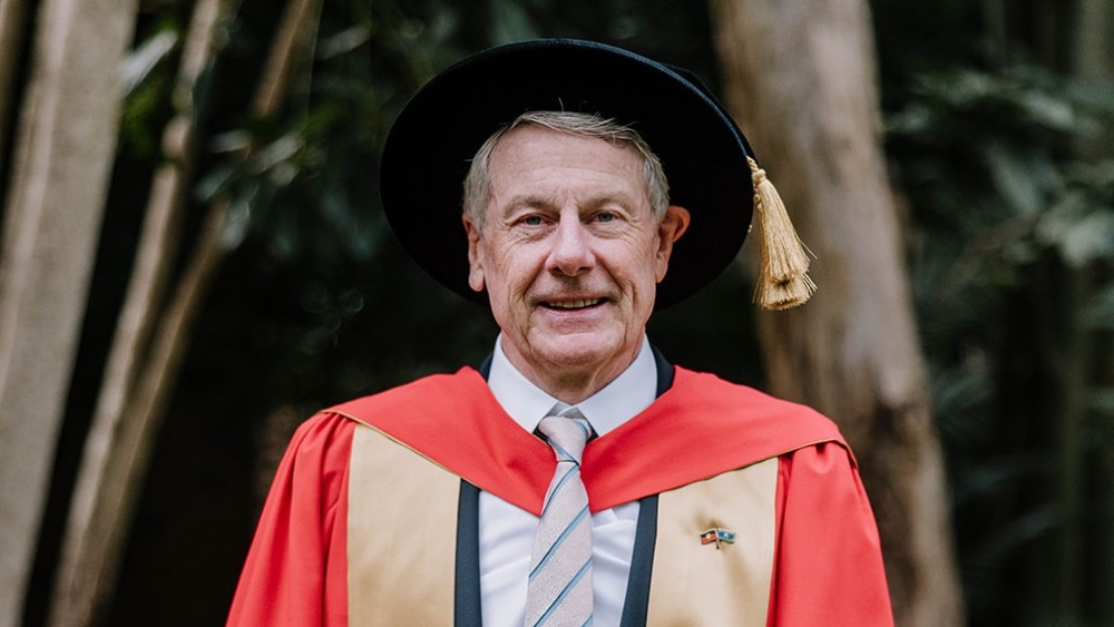 Portrait of Robert Ryan wearing red and gold academic regalia and a black velvet hat with a gold tassel. He is wearing a white shirt, a light-colored striped tie, and a small pin featuring the Aboriginal and Torres Strait Islander flags. He stands outdoors against a soft-focus background of trees.