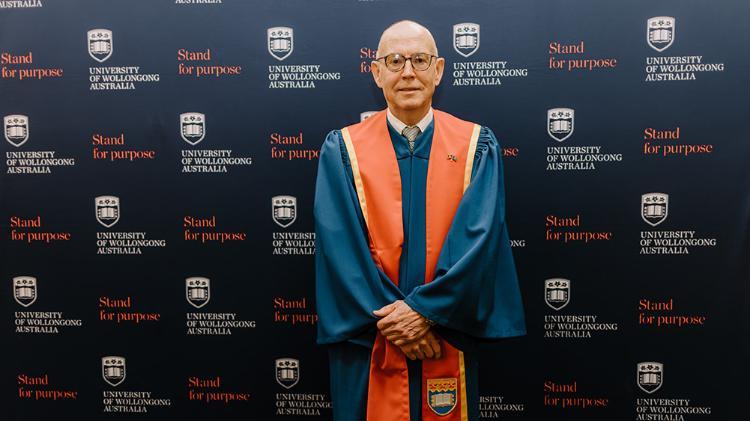 John Nowlan in Honorary Gown in front of navy wall with repeated UOW logo